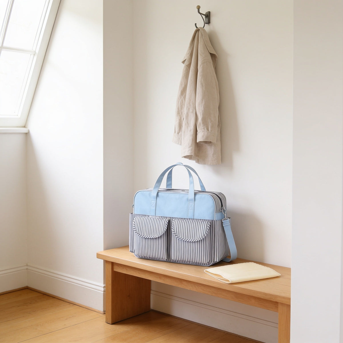 Navy and white pinstripe print diaper tote bag on a light oak hallway bench, its changing mat folded beside it.