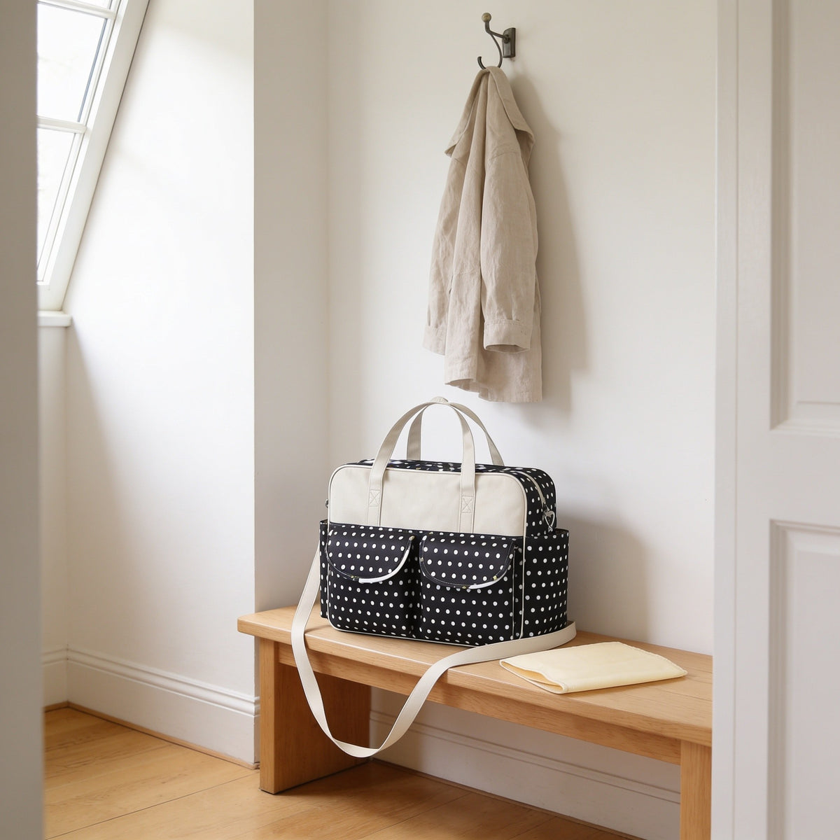 Black polka dot print diaper tote bag on a light oak hallway bench, its changing mat folded beside it.