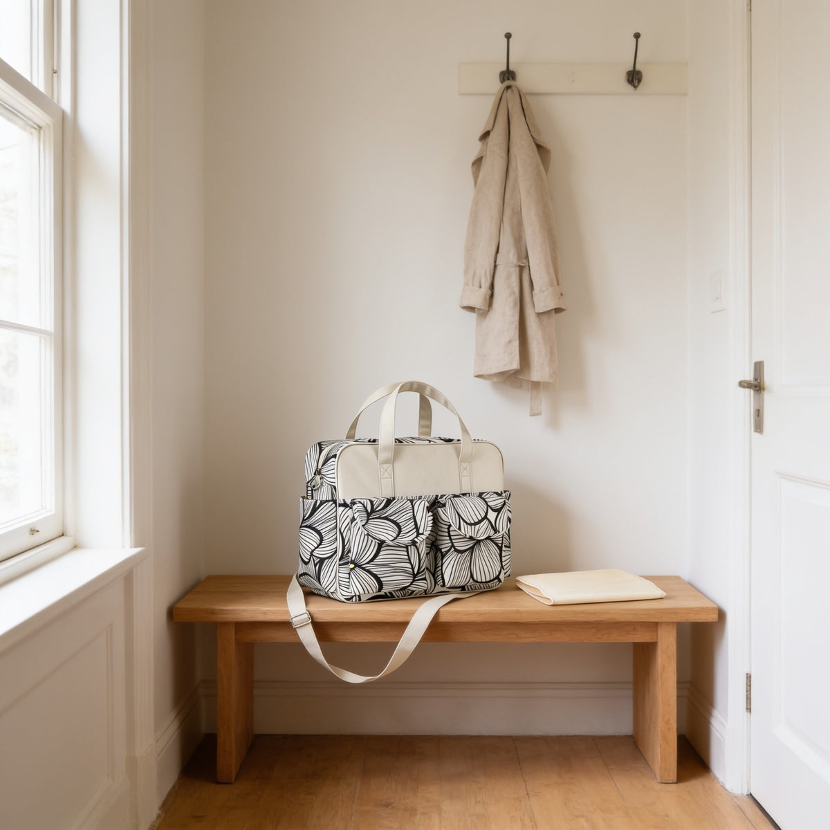 Black and white leaf print diaper tote bag on a light oak hallway bench, its changing mat folded beside it.