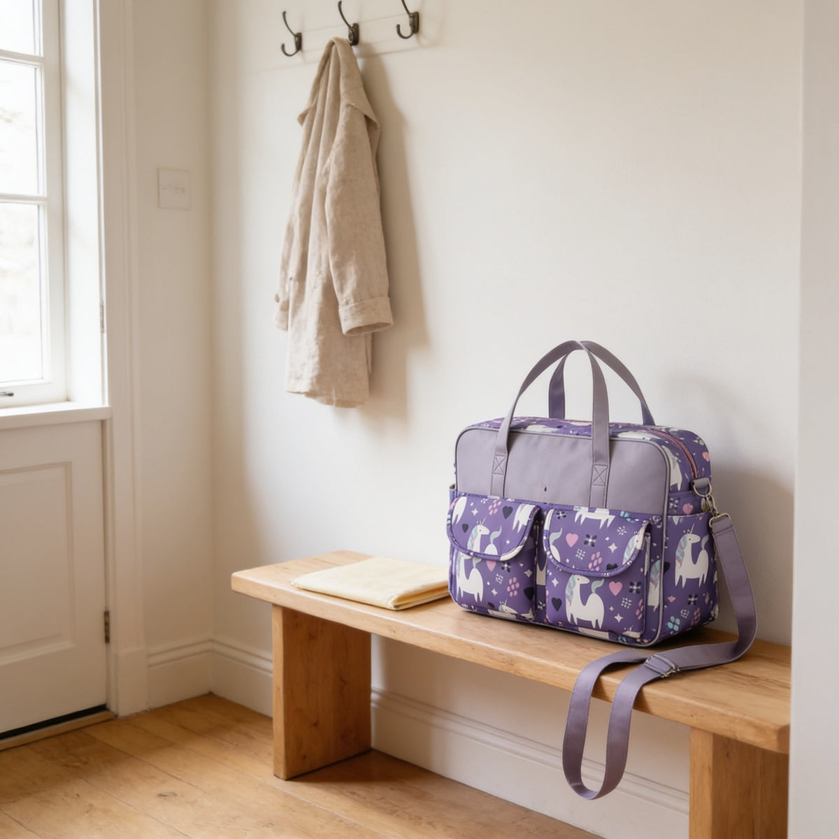 Purple unicorn print diaper tote bag on a light oak hallway bench, its changing mat folded beside it.