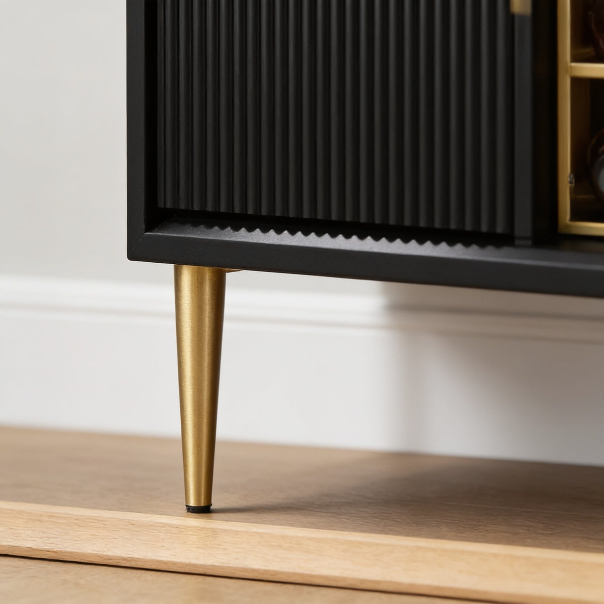Close low view of one tapered brushed gold leg lifting the fluted cabinet fourteen and a half centimetres clear of a light oak floor.